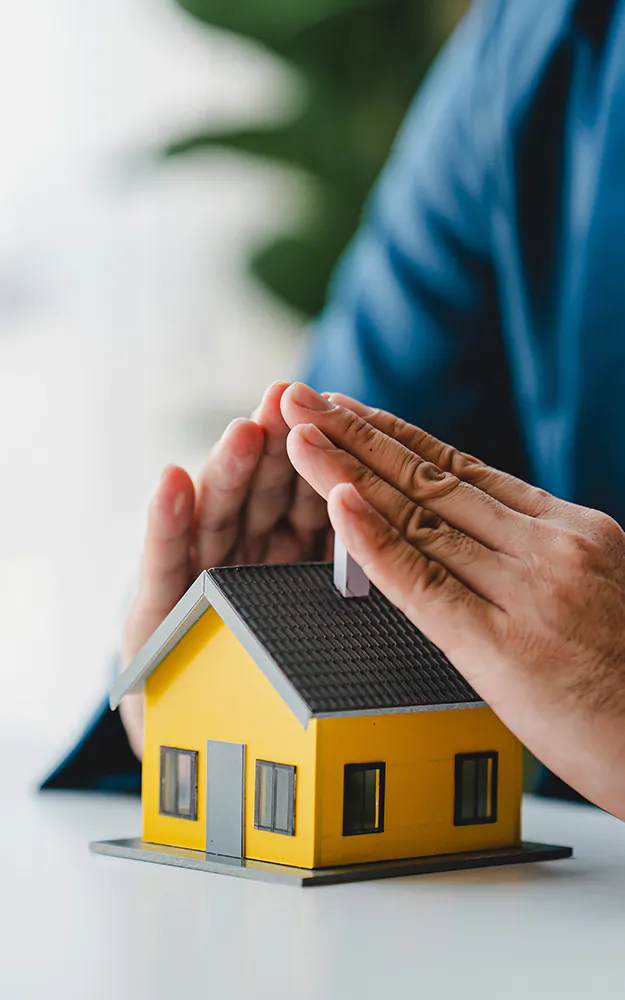 Modellino casa gialla protetto da mani a forma di tetto per tutela del patrimonio e consulenza immobiliare.